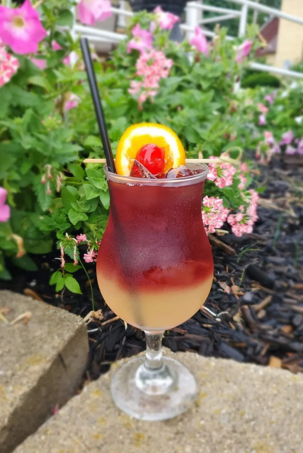 A tall cocktail garnished with orange and cherry on the patio
